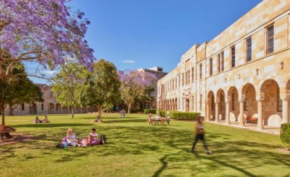 People sitting on a lawn under a jacaranda tree with sandstone buildings behind them. 